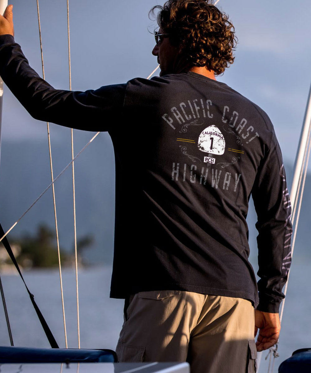 A man wearing a black long sleeve crewneck T-shirt with the text 'PACIFIC COAST HIGHWAY' on it, standing near water. View 2