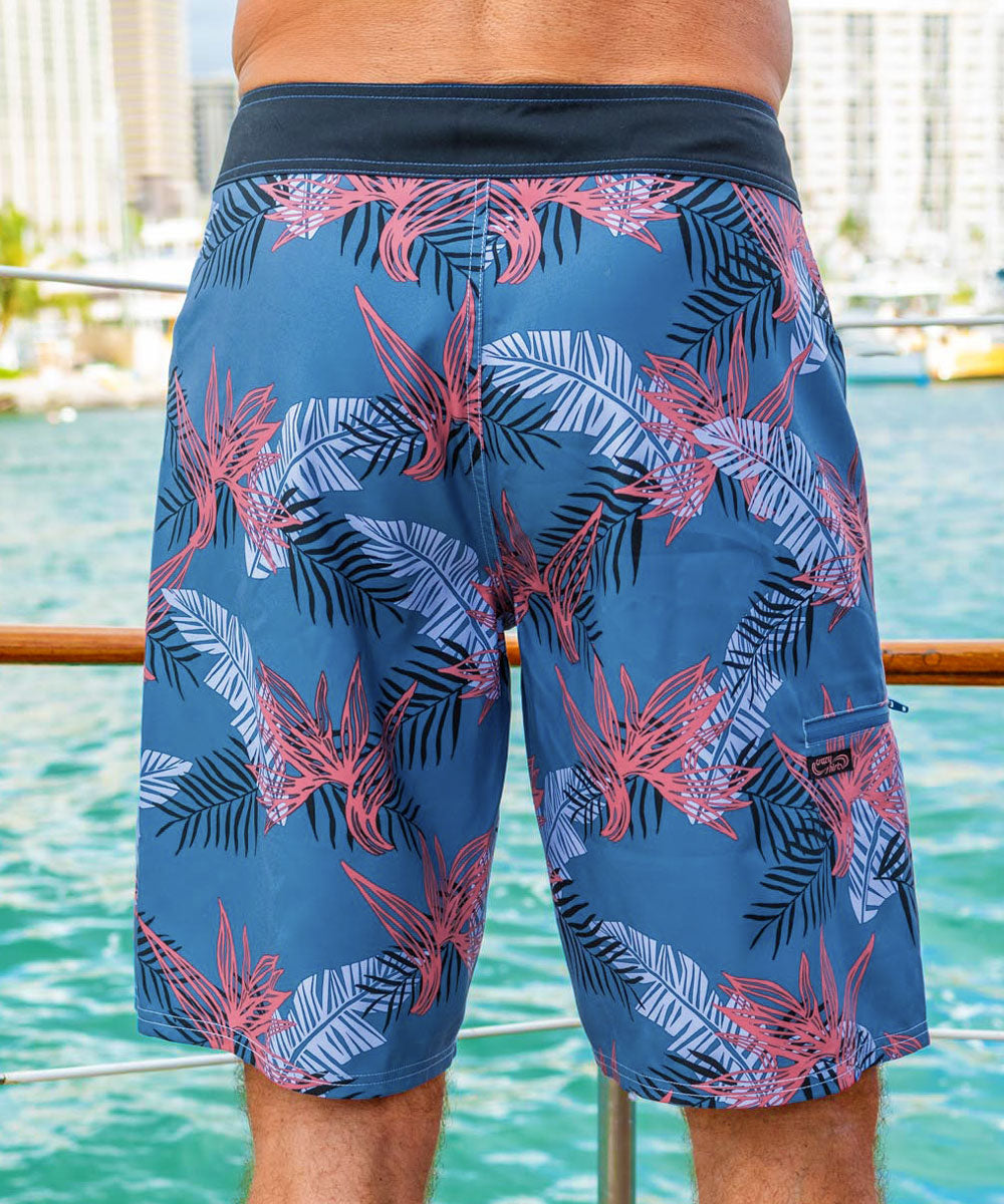 A person wearing a pair of slate blue board shorts with a tribal pattern, displayed in a beach setting, facing away from the camera. View 3