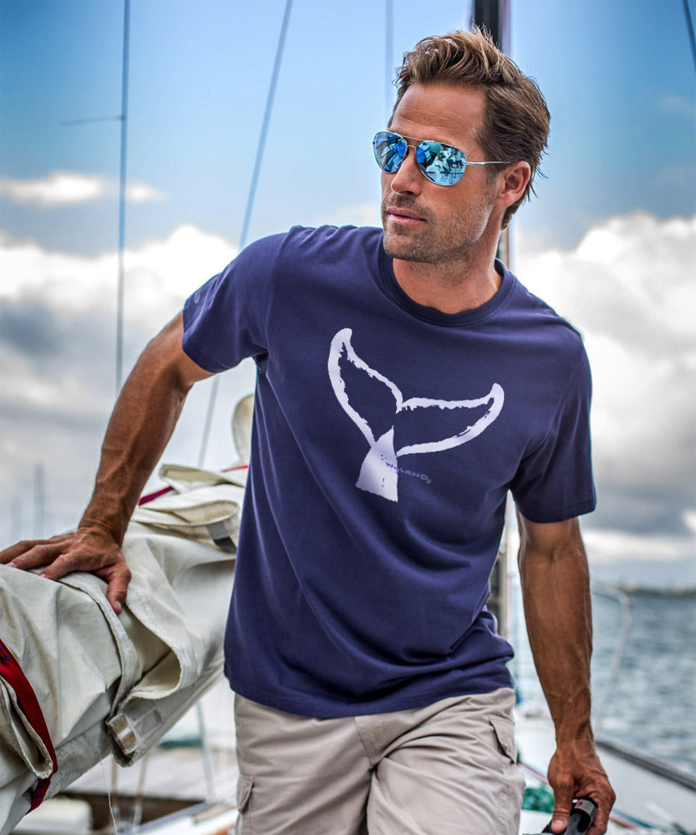 A man wearing an indigo blue short sleeve Pima cotton T-shirt with a whale tail graphic design, standing on a boat. View 2