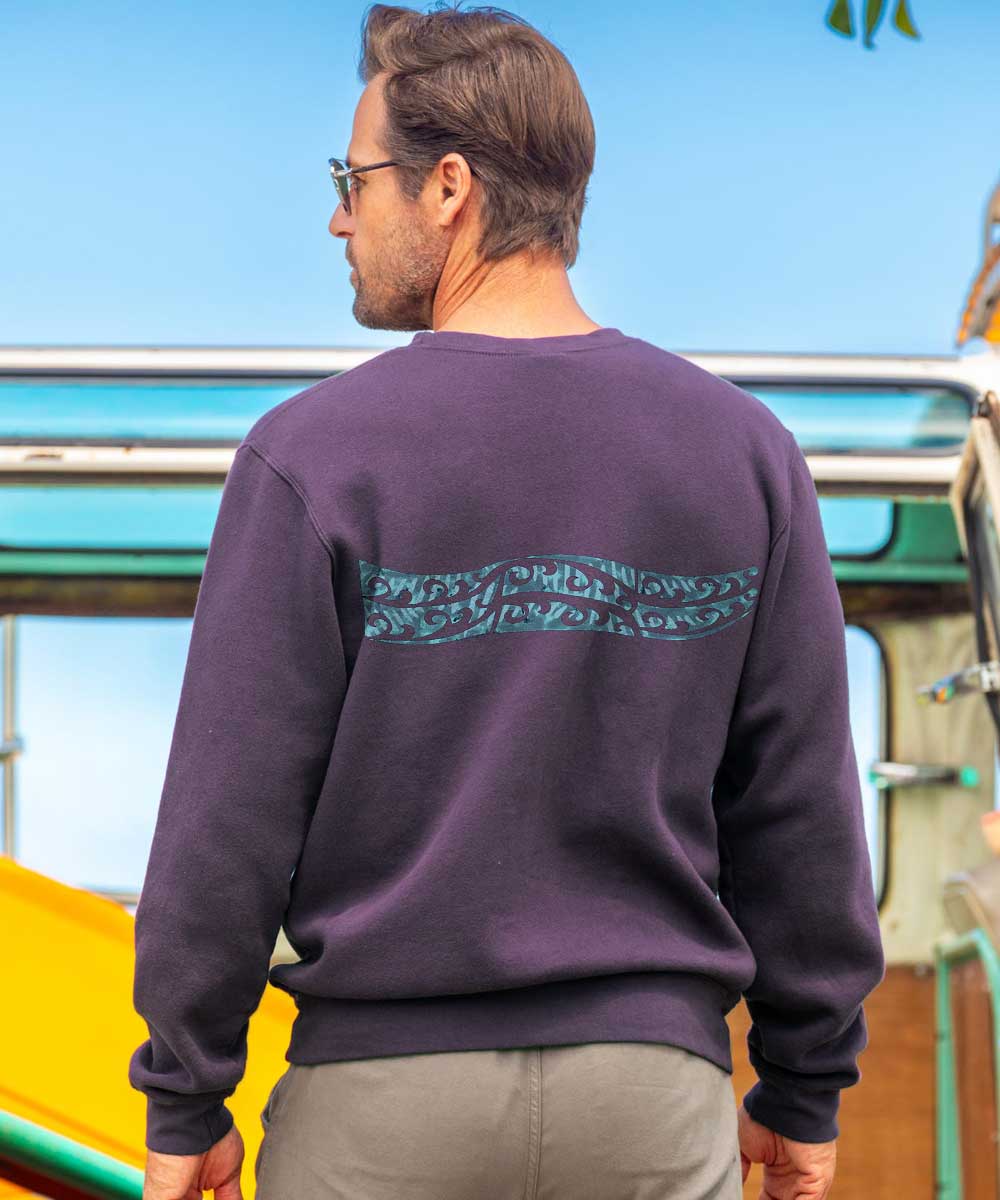A man facing away from the camera wearing a purple long sleeve with a blue tribal design going across the back, standing near a vintage van. View 3