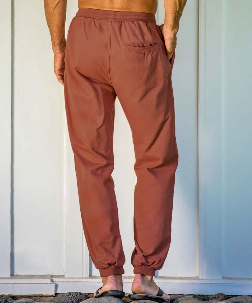 A person standing by a light blue wall wearing rust pants and black slippers, with hands in pockets and showcasing the back right pocket. View 3