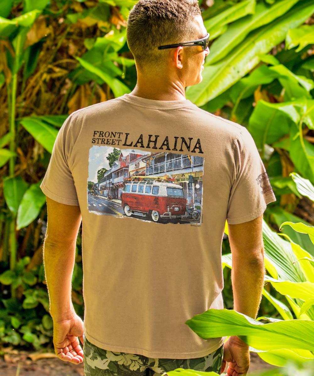 A person wearing a beige crewneck t-shirt with a graphic of a Volkswagen van on the front, standing among green foliage. View 2