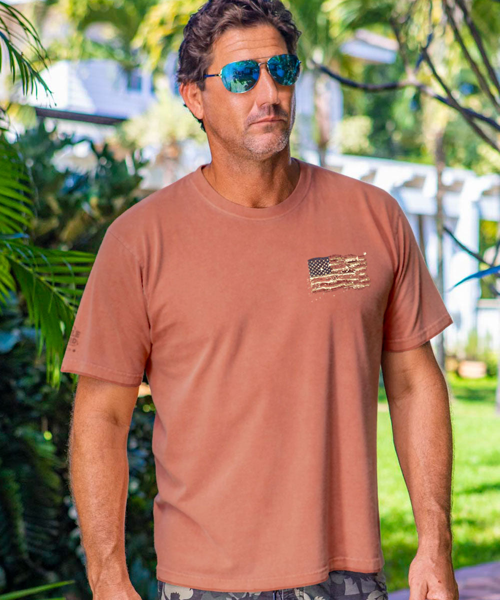 Man wearing a light red shirt with a small logo on chest with greenery surrounding. View 3
