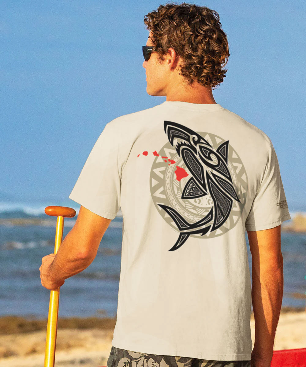 A person standing on a beach wearing a beige short-sleeve crewneck t-shirt with a tribal shark design on the back. View 2