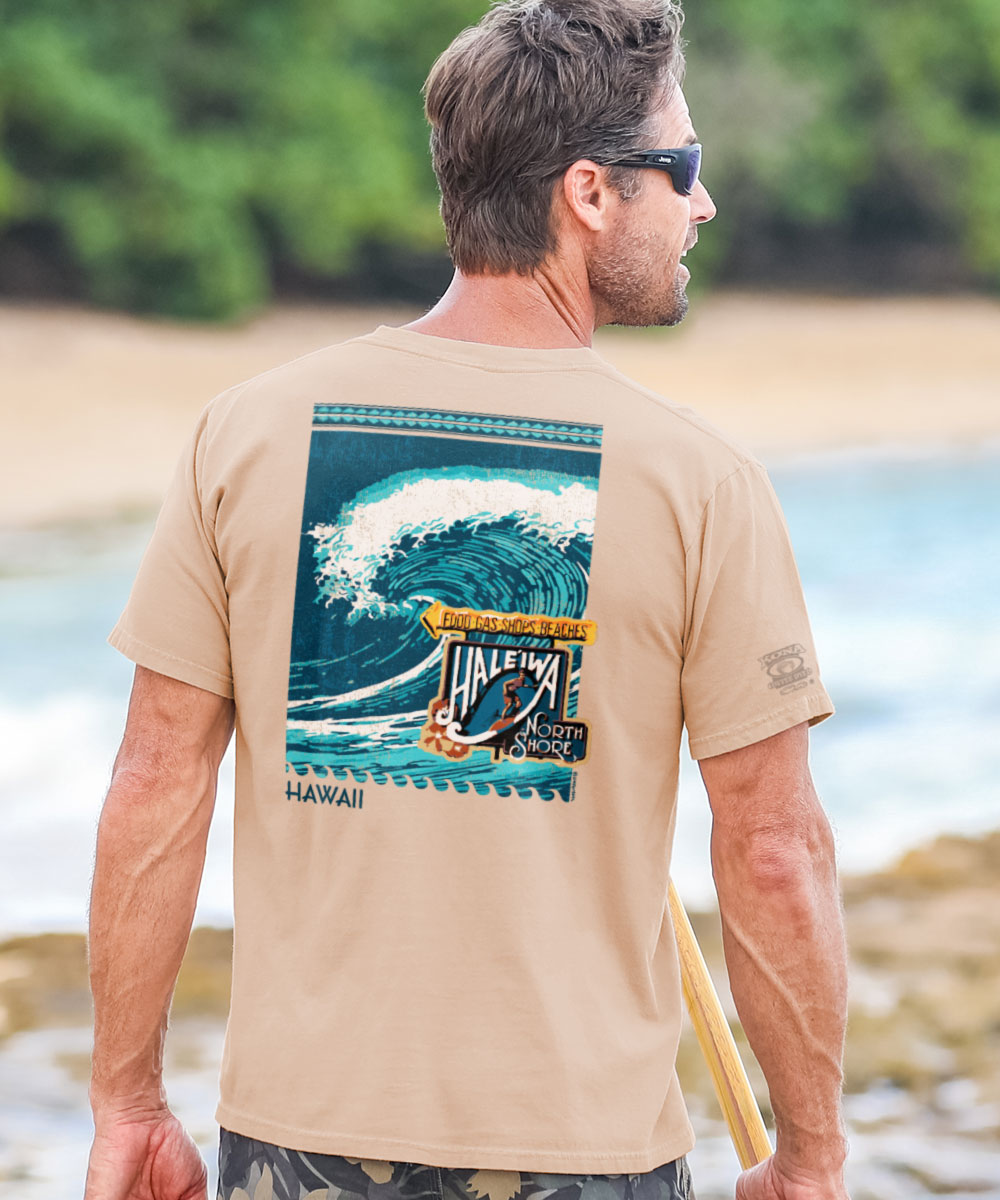 A man wearing a beige short sleeve crewneck t-shirt with a graphic of a wave and the Haleiwa sign in front of a beach setting. View 2