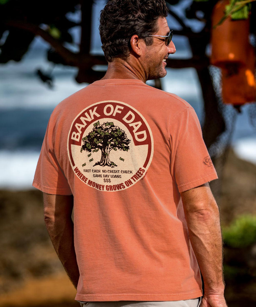 A man wearing a salmon short sleeve crewneck t-shirt with the text '_BANK OF Dad' printed on the back, featuring a tree illustration. View 2