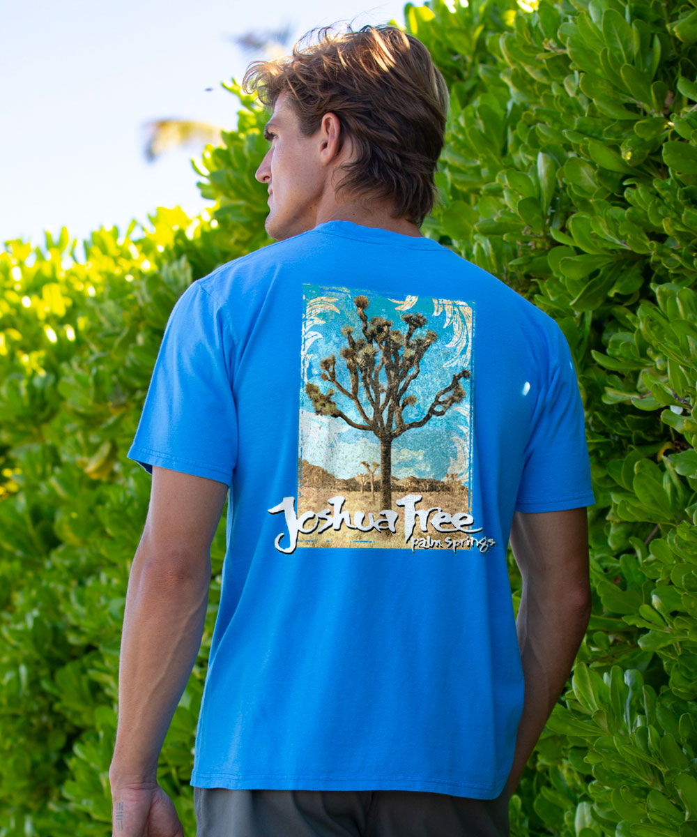 Person wearing a blue t-shirt with a Joshua Tree design, standing in front of green foliage. View 2