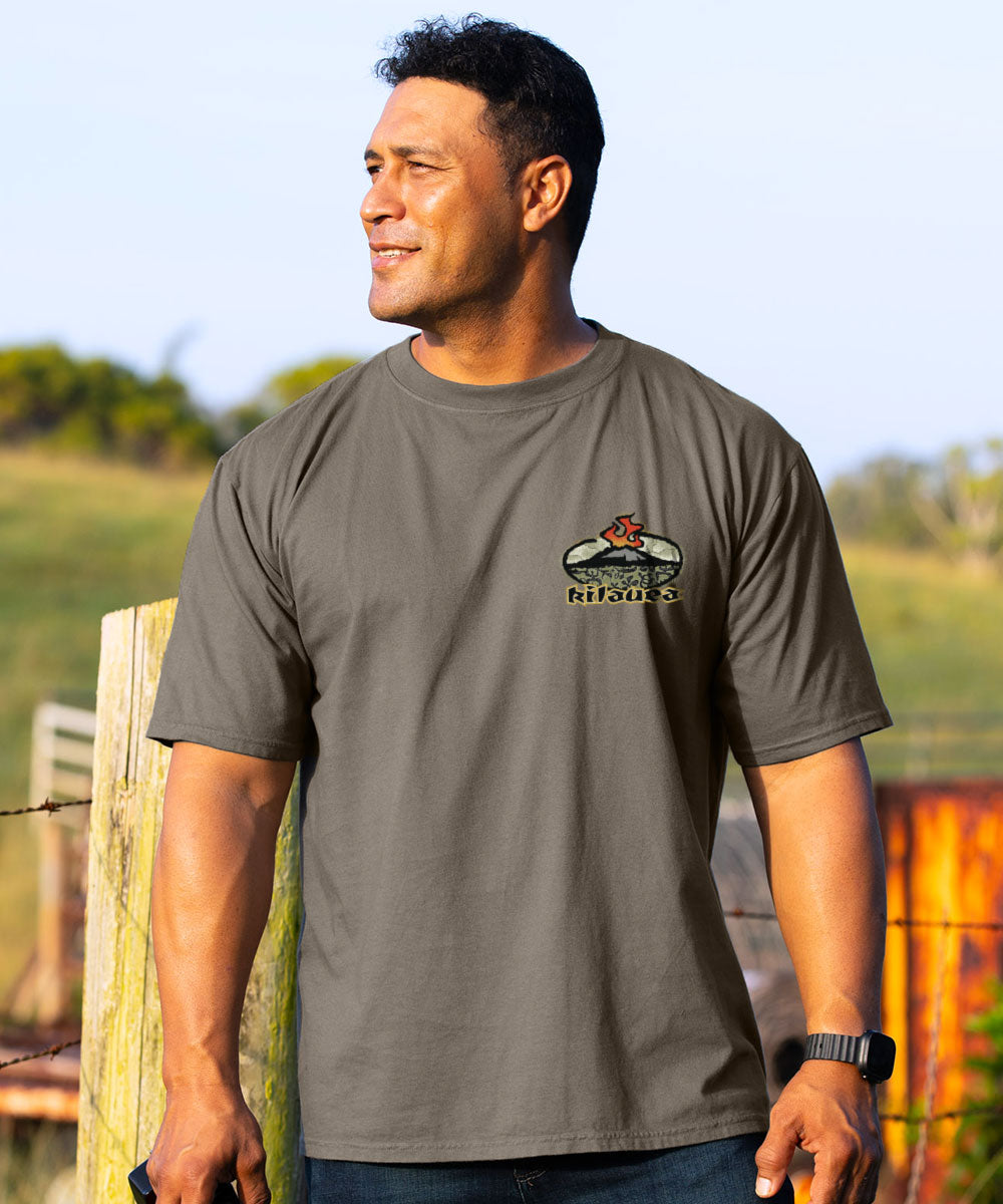 A man wearing a dark gray crewneck t-shirt with a small 'Rustols' logo on the left chest, standing outdoors in front of a rustic fence. View 3
