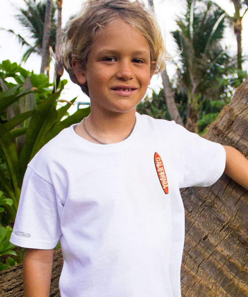 Child wearing a white t-shirt with a logo, standing next to a palm tree in a tropical setting. View 3