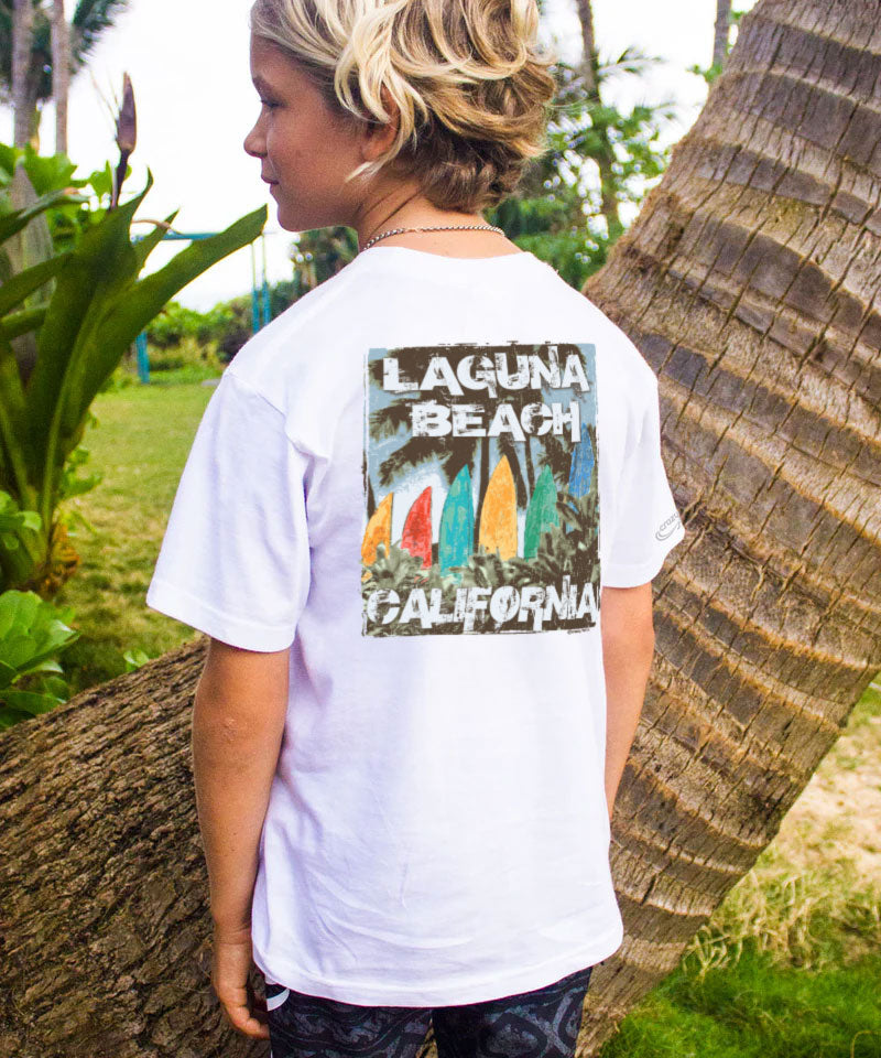 Person wearing a white t-shirt with 'Laguna Beach, California' print leaning against a palm tree. View 2