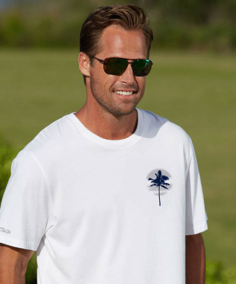 A smiling man wearing dark sunglasses and a white short-sleeve t-shirt with a small dark palm tree graphic on the left chest, set against a blurred green outdoor background. View 3