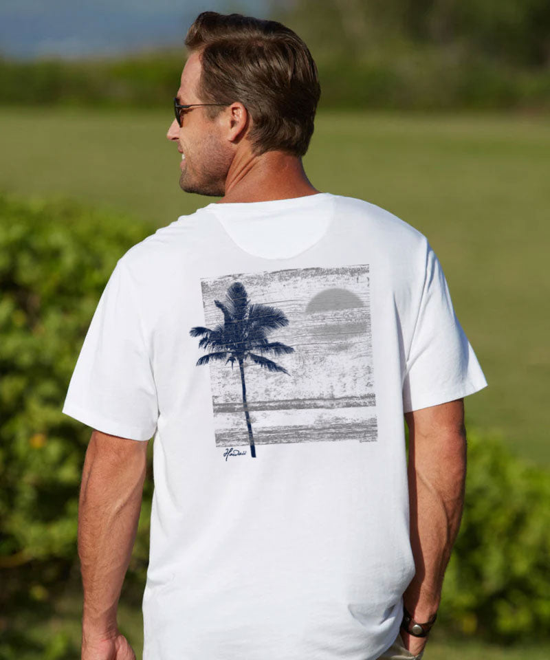 A man wearing a white short-sleeve t-shirt with a blue and gray graphic print of a palm tree and sunset on the back, photographed outdoors on a sunny day. View 2