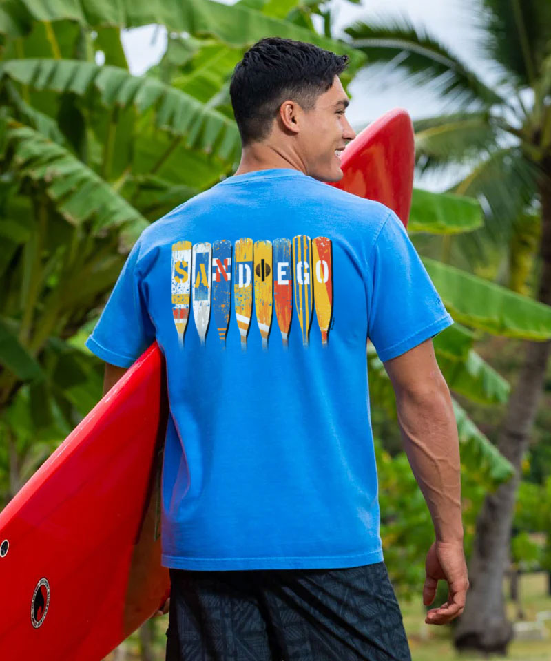 Man holding a red kayak with 'San Diego' design on a blue shirt, surrounded by green foliage. View 2