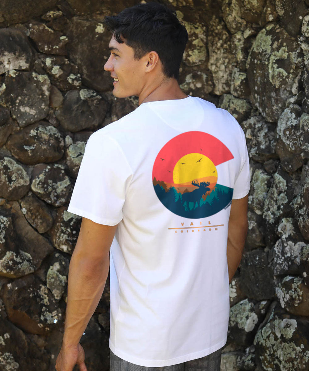 Person wearing a white t-shirt with a colorful graphic design in front of a stone wall. View 2