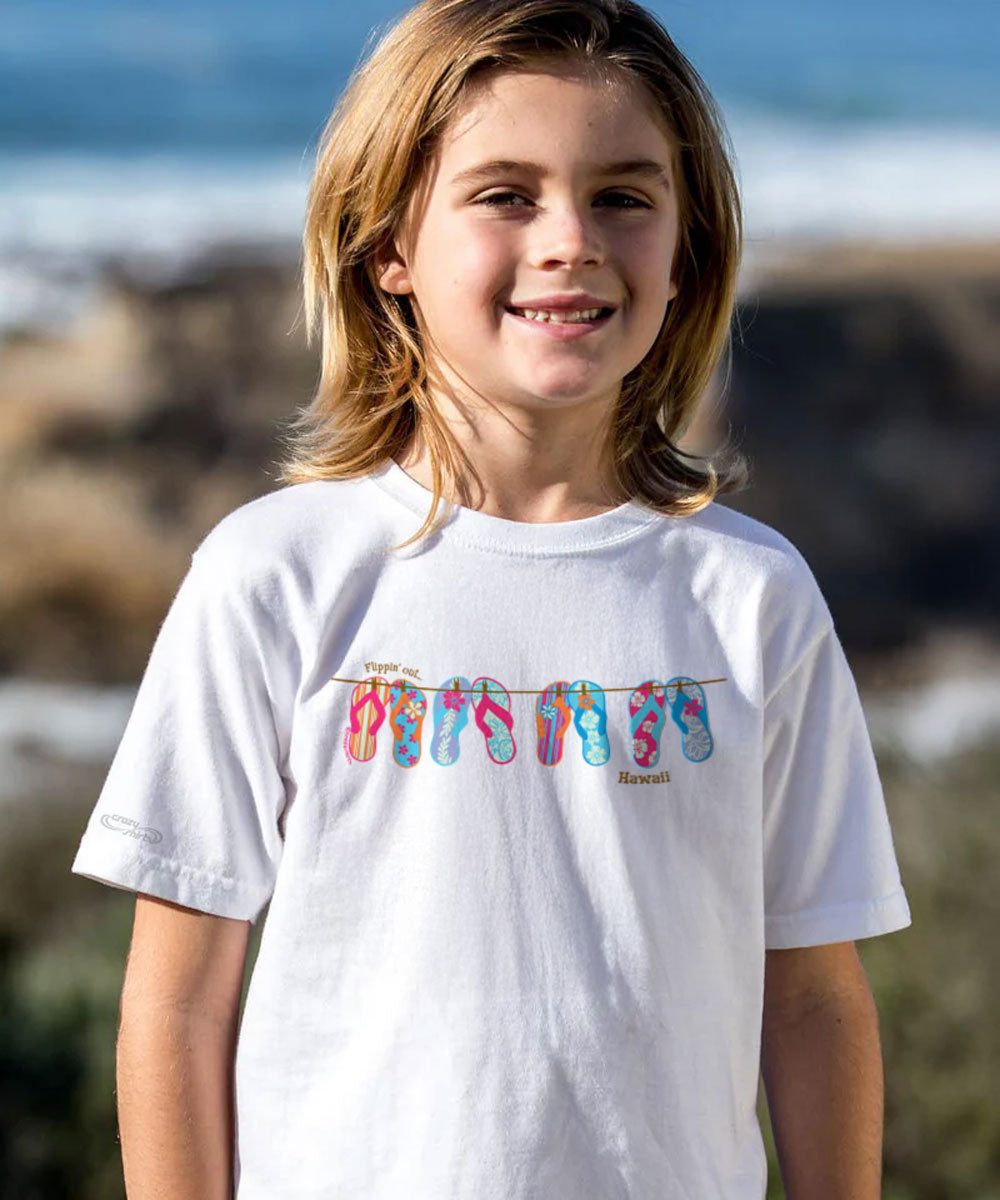 Child wearing a white t-shirt with colorful flip-flop design on a beach. View 2