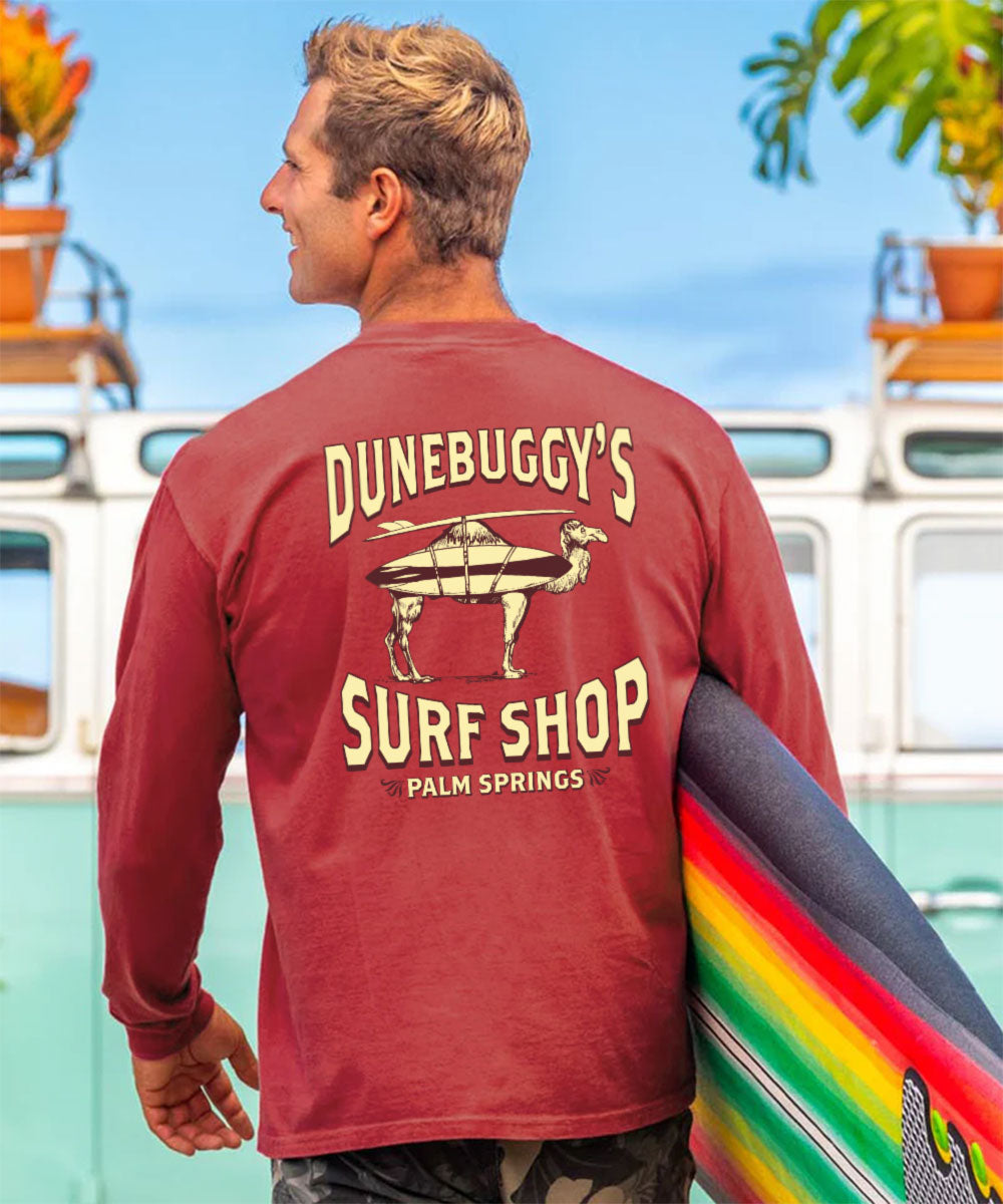 Man wearing a red 'Dunebuggy's Surf Shop' shirt holding a rainbow surfboard with a vintage van and plants in the background. View 2