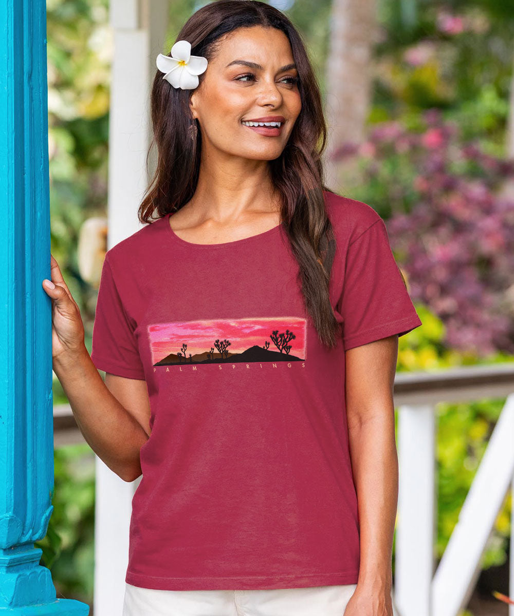 Woman wearing a maroon t-shirt with a graphic design, standing outdoors with greenery in the background. View 2