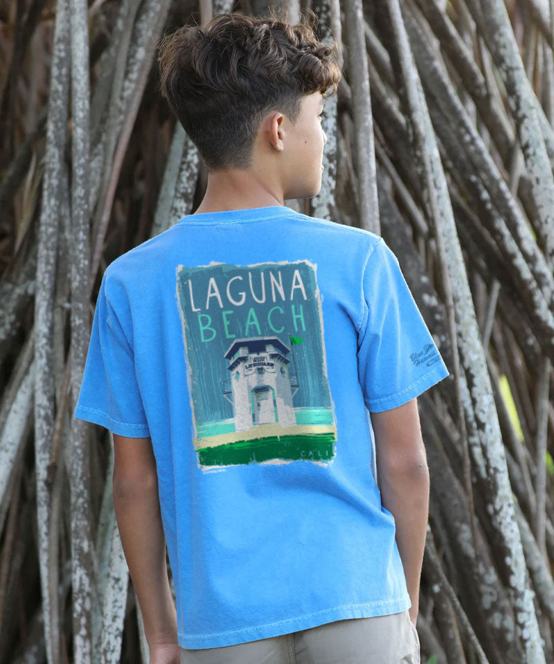 Young boy wearing a blue t-shirt with 'Laguna Beach' design, standing in front of tree trunks. View 2