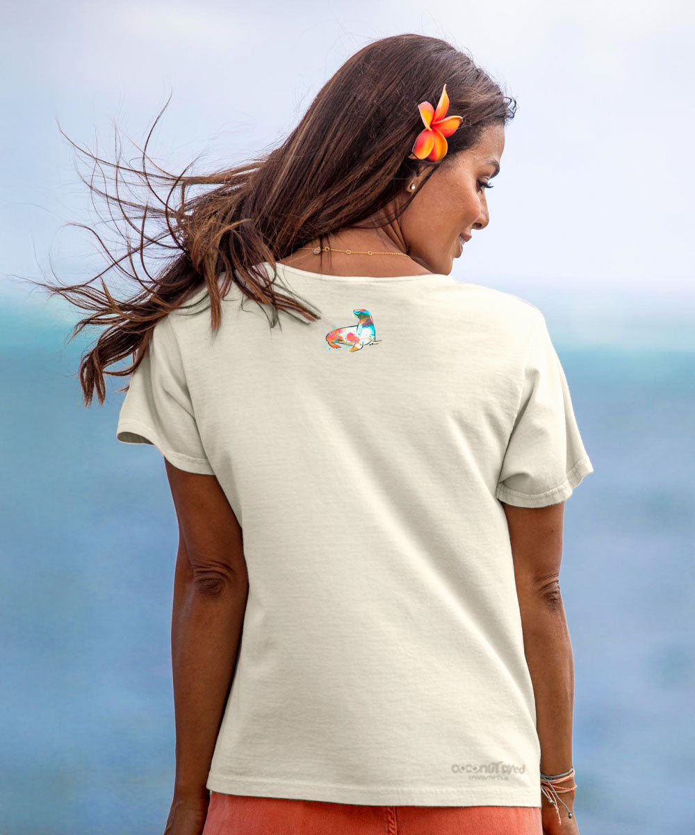 Woman wearing a beige t-shirt with a small design, standing against a blurred natural background. View 3
