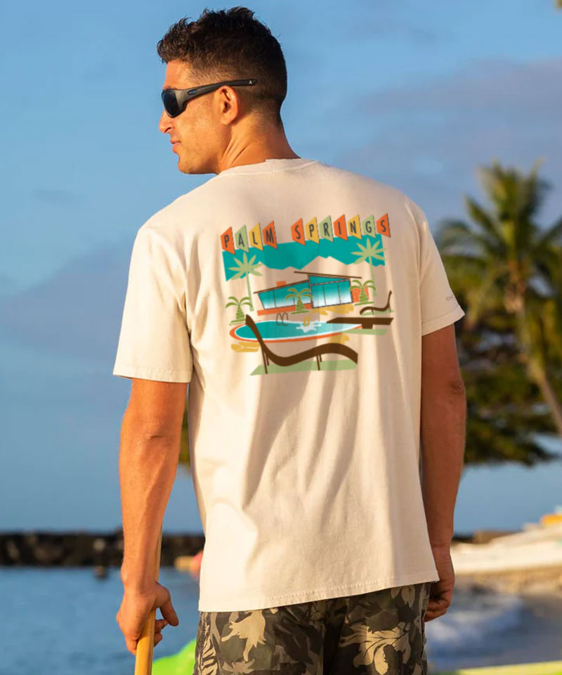 Man wearing a t-shirt with a colorful graphic on the back, standing by a body of water with palm trees. View 2