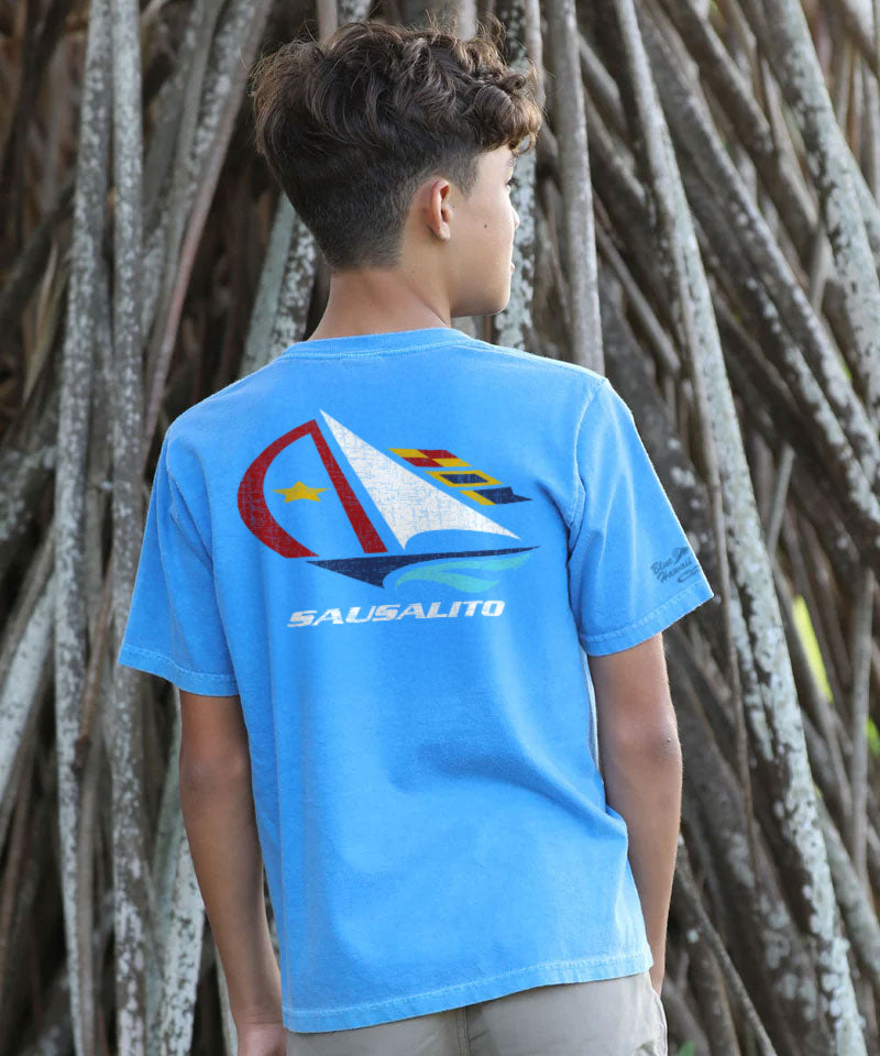 Person wearing a blue t-shirt with a sailboat design and 'Sausalito' text, standing against a textured wall. View 2