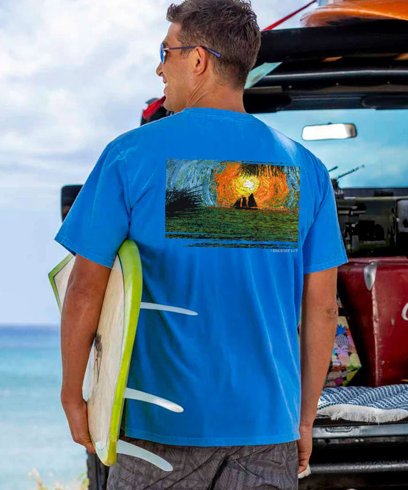 Man wearing a blue shirt with a sunset ocean design holding a surfboard looking at the ocean View 2