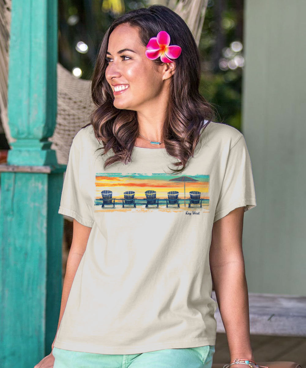 Woman wearing a beige t-shirt with a beach chair design across the chest, standing in front of a green wall. View 2