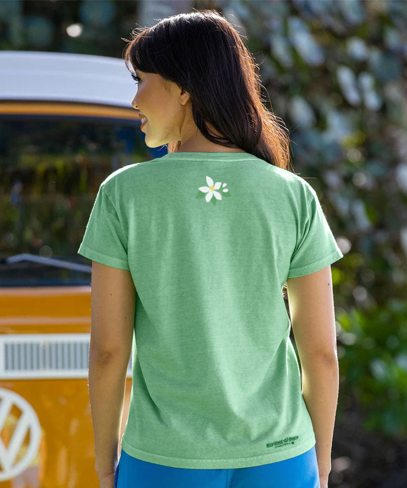 Woman facing away from camera, wearing a light green t-shirt with a small floral logo on top middle, standing outdoors. View 3