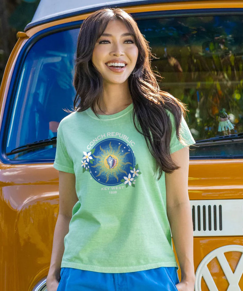 Woman wearing a light green t-shirt with a graphic 'Conch Republic Key West 1828' design and floral aspects on center chest  in front of an orange van. View 2