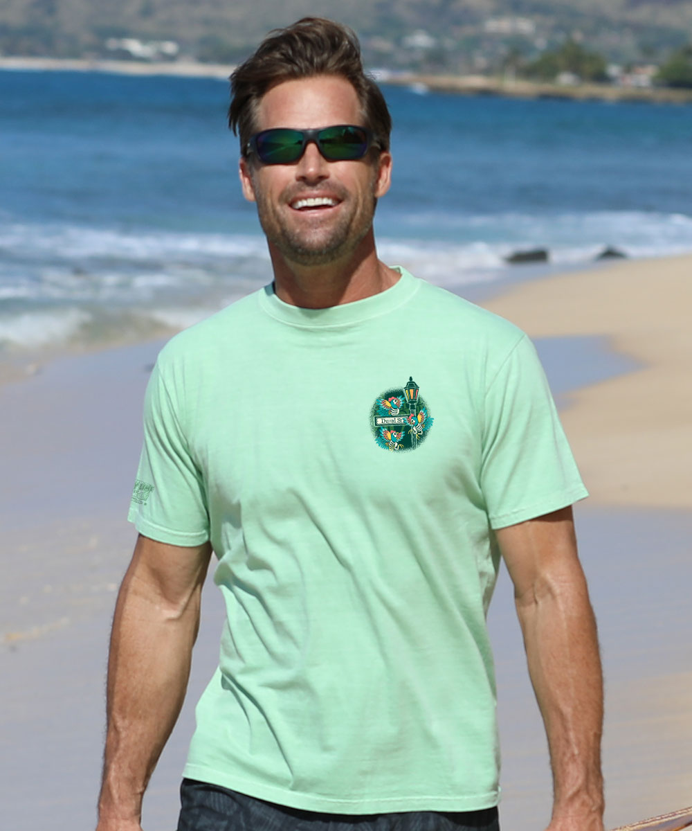 A man wearing a light mint green t-shirt with a circular green and blue graphic on the left chest, standing on a sandy beach with the ocean in the background. View 3