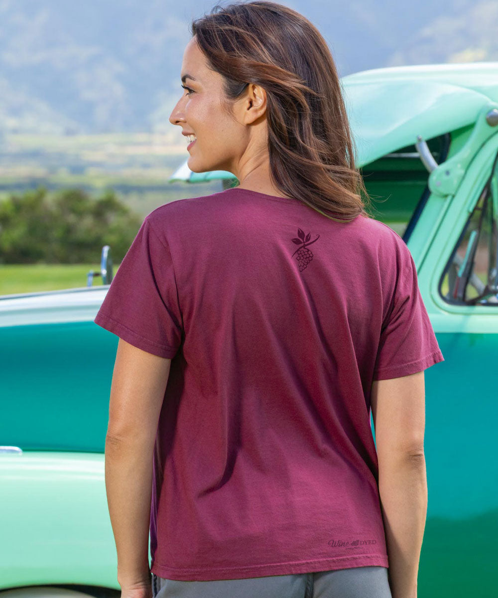 Woman wearing a maroon t-shirt with a logo, standing in front of a vintage green truck. View 3