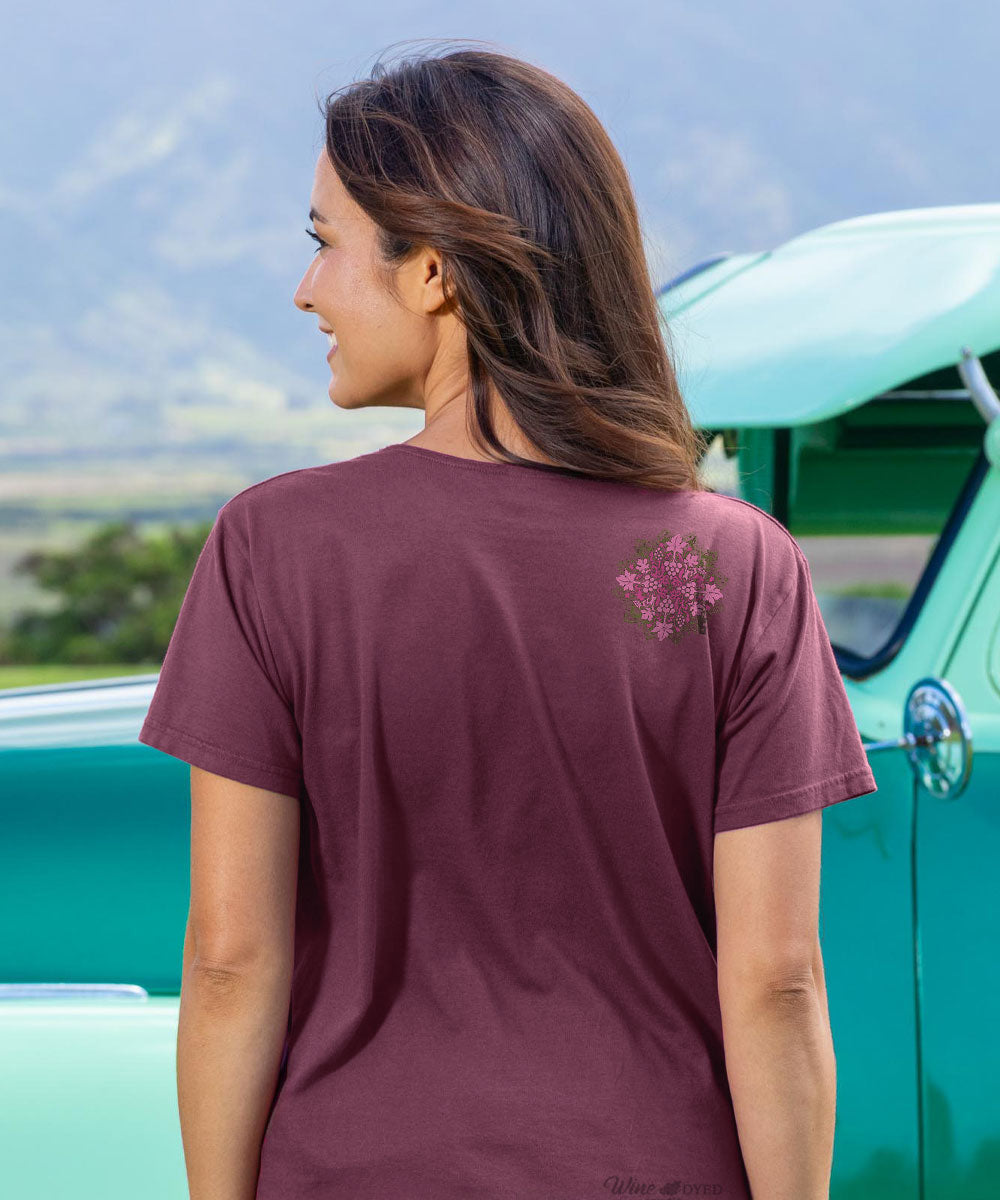 Woman wearing a maroon t-shirt with a floral design, standing in front of a turquoise vintage car. View 3
