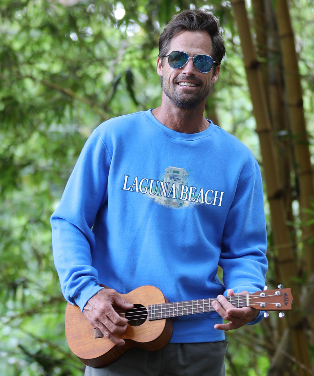 Man wearing a blue 'Laguna Beach' sweatshirt holding a ukulele with a green, leafy background View 2