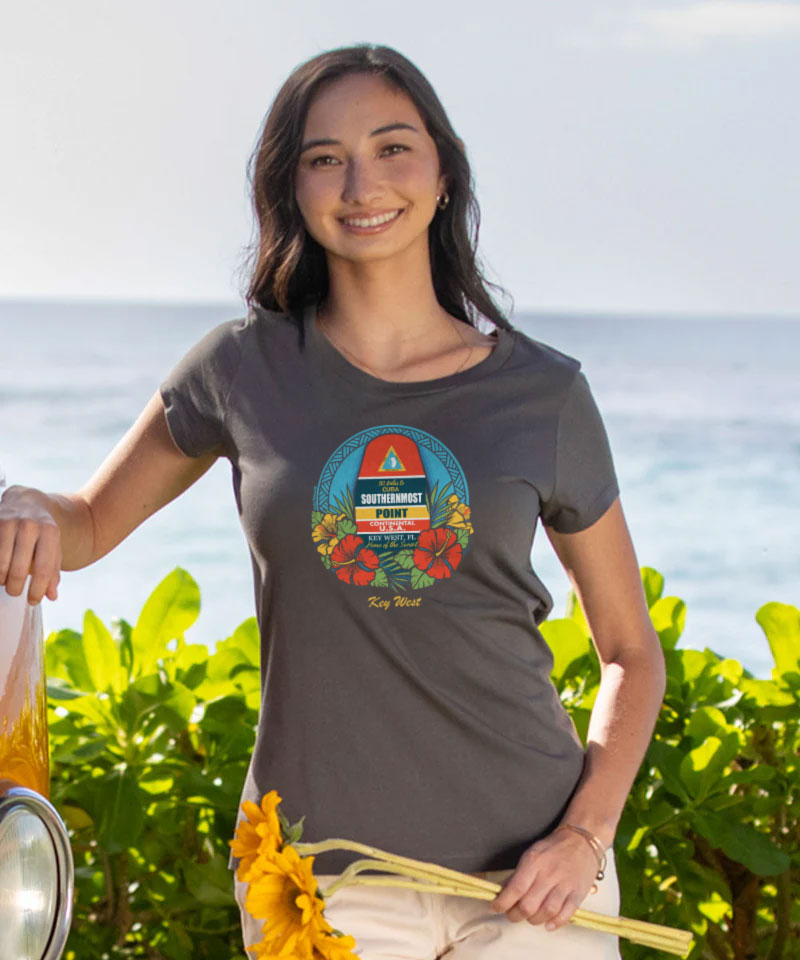 Woman wearing a gray t-shirt with a colorful graphic design, standing outdoors with greenery and water in the background. View 2