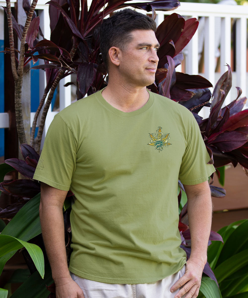 A man wearing a light olive green crewneck t-shirt with a small embroidered green and gold eagle logo on the left chest, standing in front of tropical plants. View 3