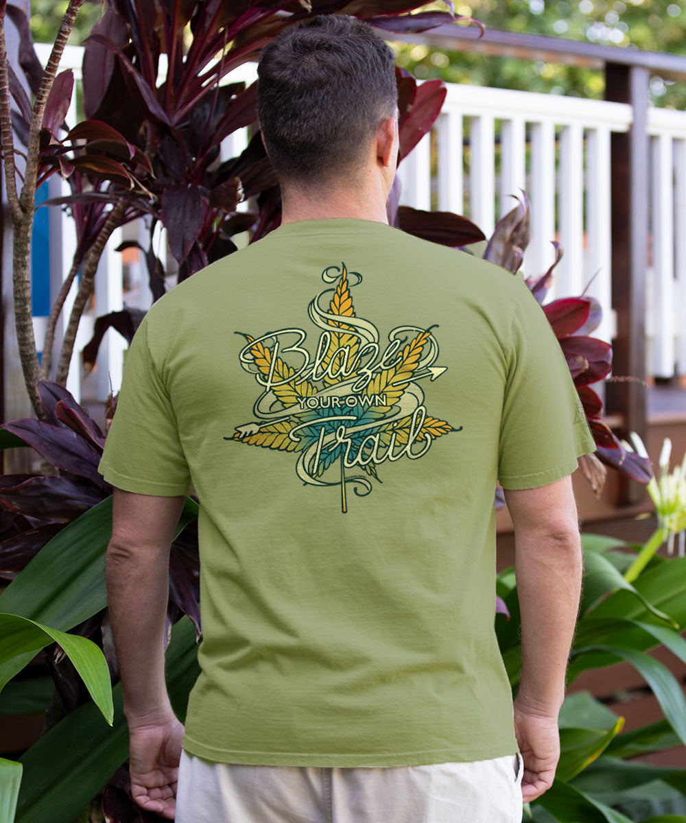 A man viewed from behind wearing an olive green t-shirt with a large graphic on the back that reads 'Back to the Own Trail' in yellow and teal. He is standing outdoors in front of tropical plants. View 2