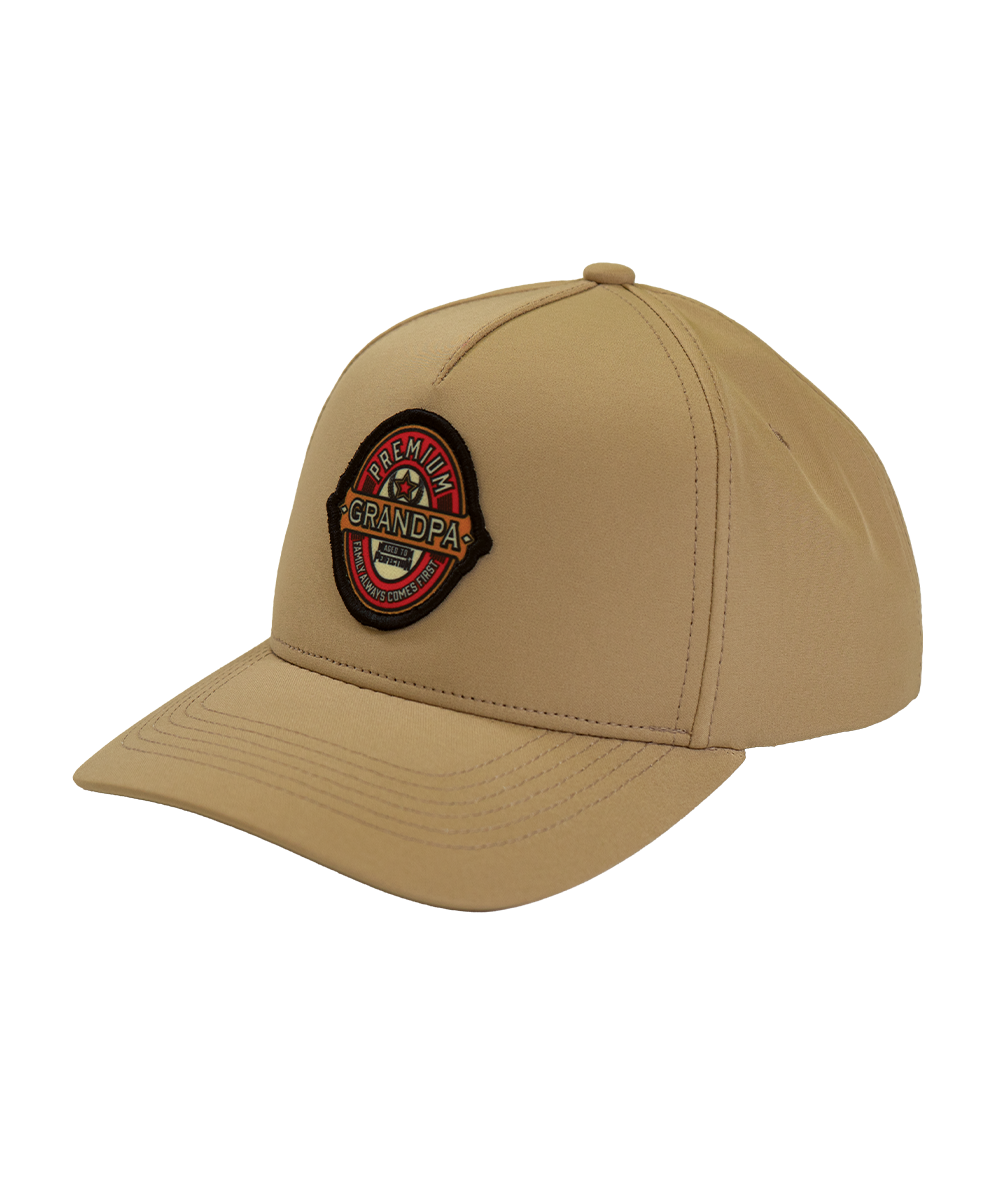 A khaki beige baseball cap featuring a circular embroidered patch on the front that reads 'PREMIUM GRANDPA' with a star and anchor design. View 1