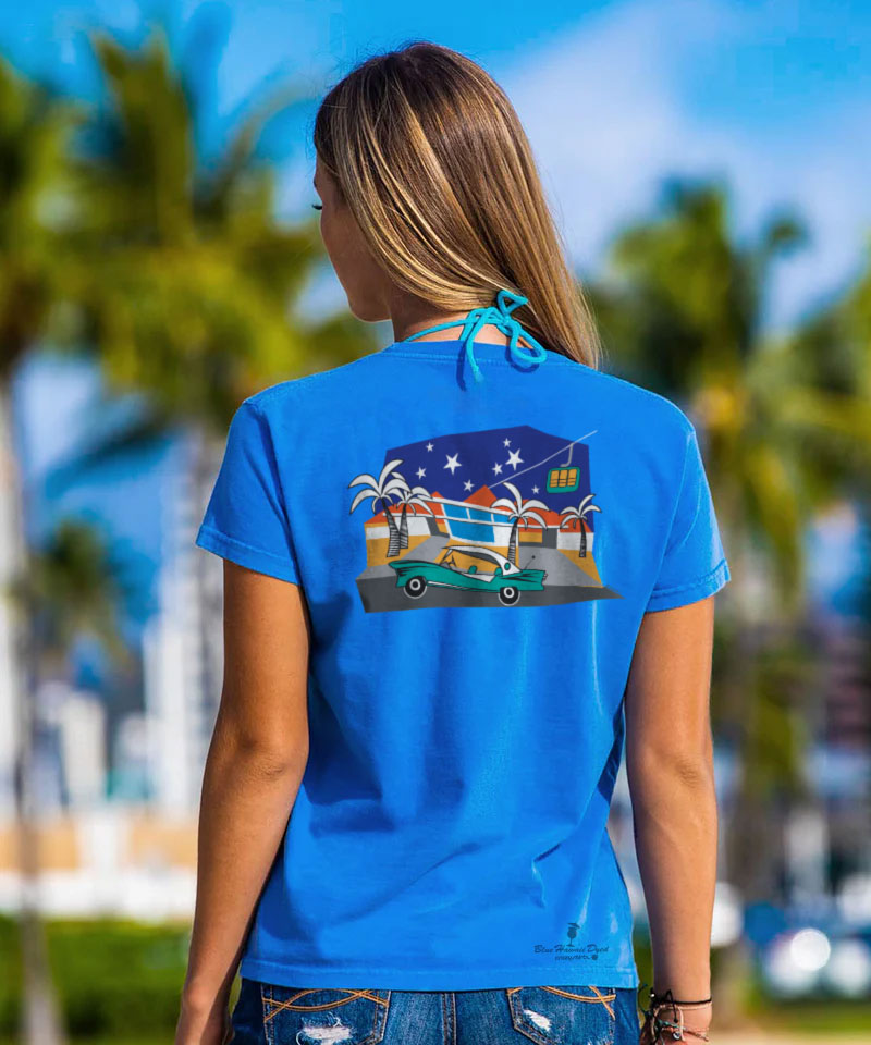 Woman wearing a blue t-shirt with a graphic design of a beach scene on the back, standing outdoors with palm trees in the background. View 2