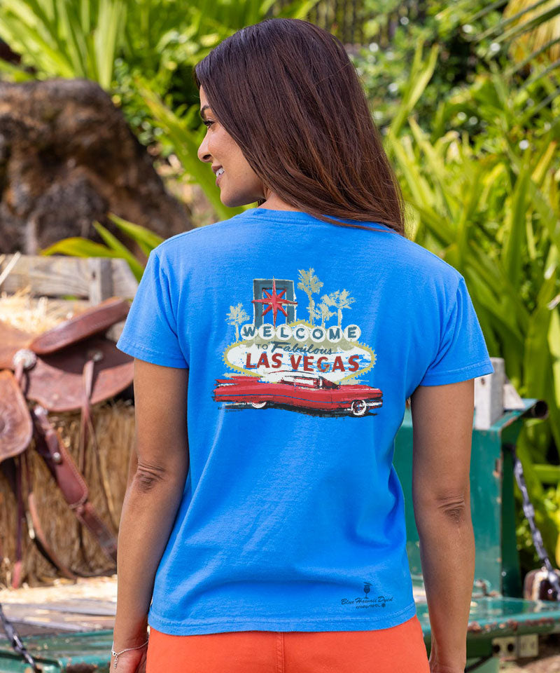 Woman wearing a blue t-shirt with Las Vegas graphic, standing outdoors with greenery in the background View 3