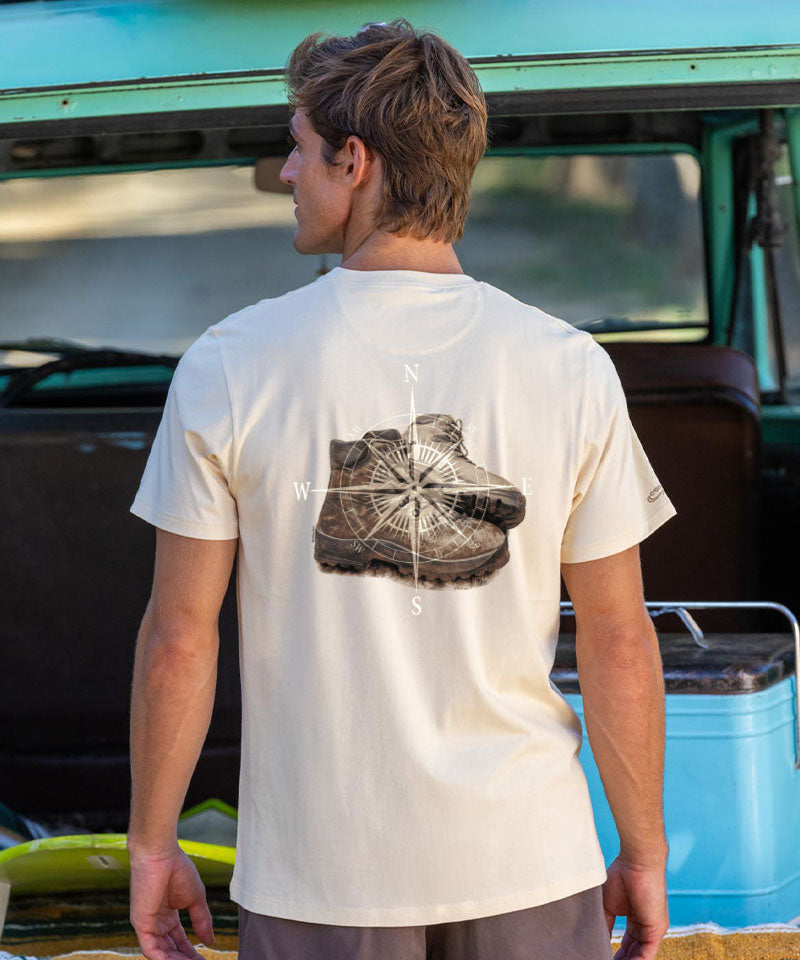Person wearing a beige t-shirt with a graphic design of shoes and compass directions, standing in front of a vintage van. View 2
