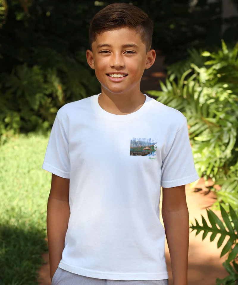 Young boy wearing a white t-shirt with a small graphic on it, standing outdoors with greenery in the background. View 3