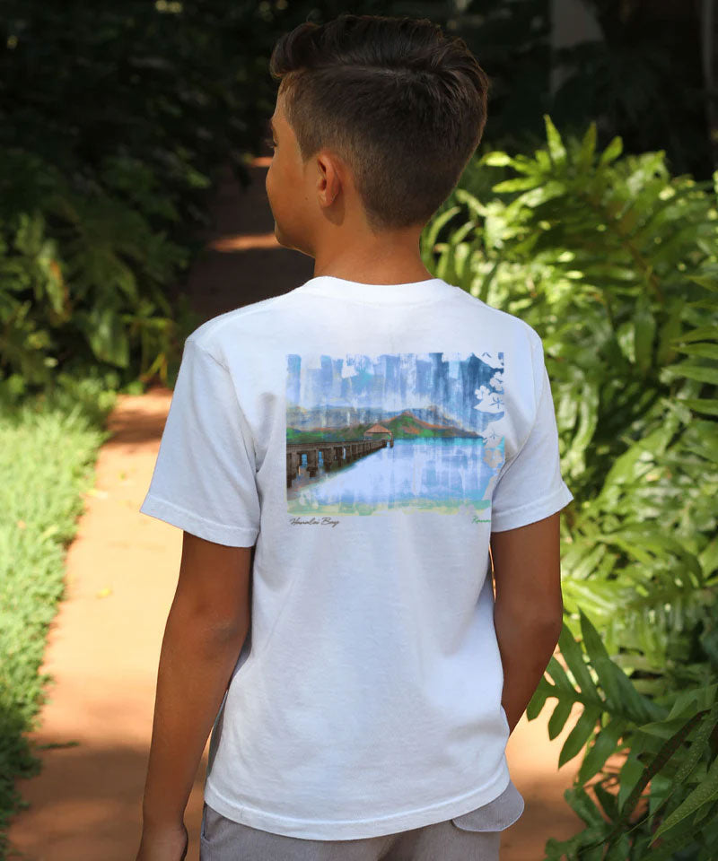 Child wearing a white t-shirt with a scenic design, standing outdoors among greenery. View 2