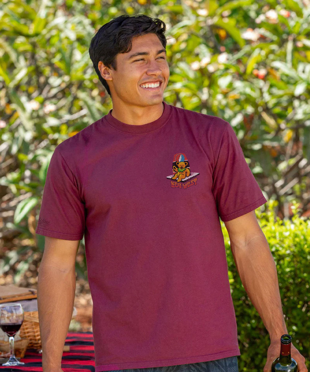 Man wearing a maroon t-shirt with a logo, standing outdoors with greenery in the background. View 3
