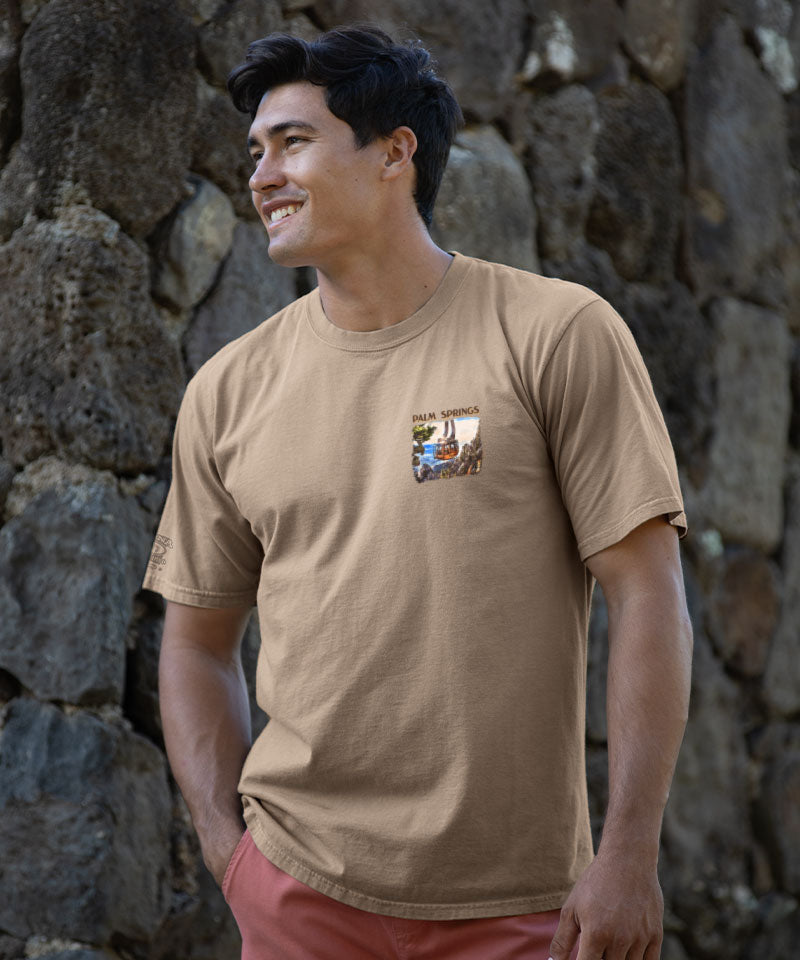 Man wearing a light brown t-shirt with a graphic design standing against a stone wall. View 3