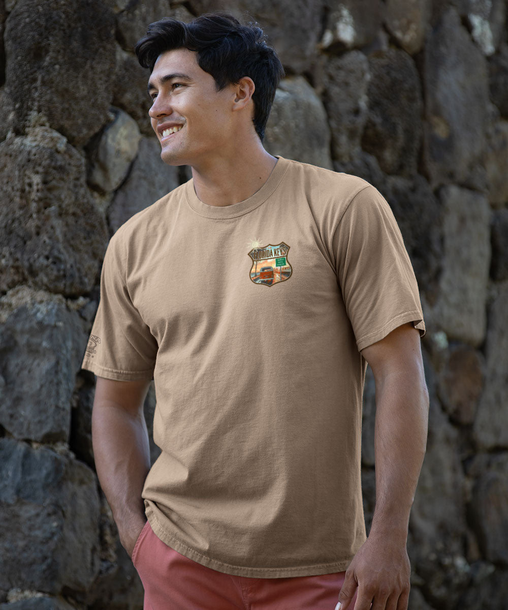 Man wearing a light brown t-shirt with a small logo on the front, standing against a stone wall.  View 3