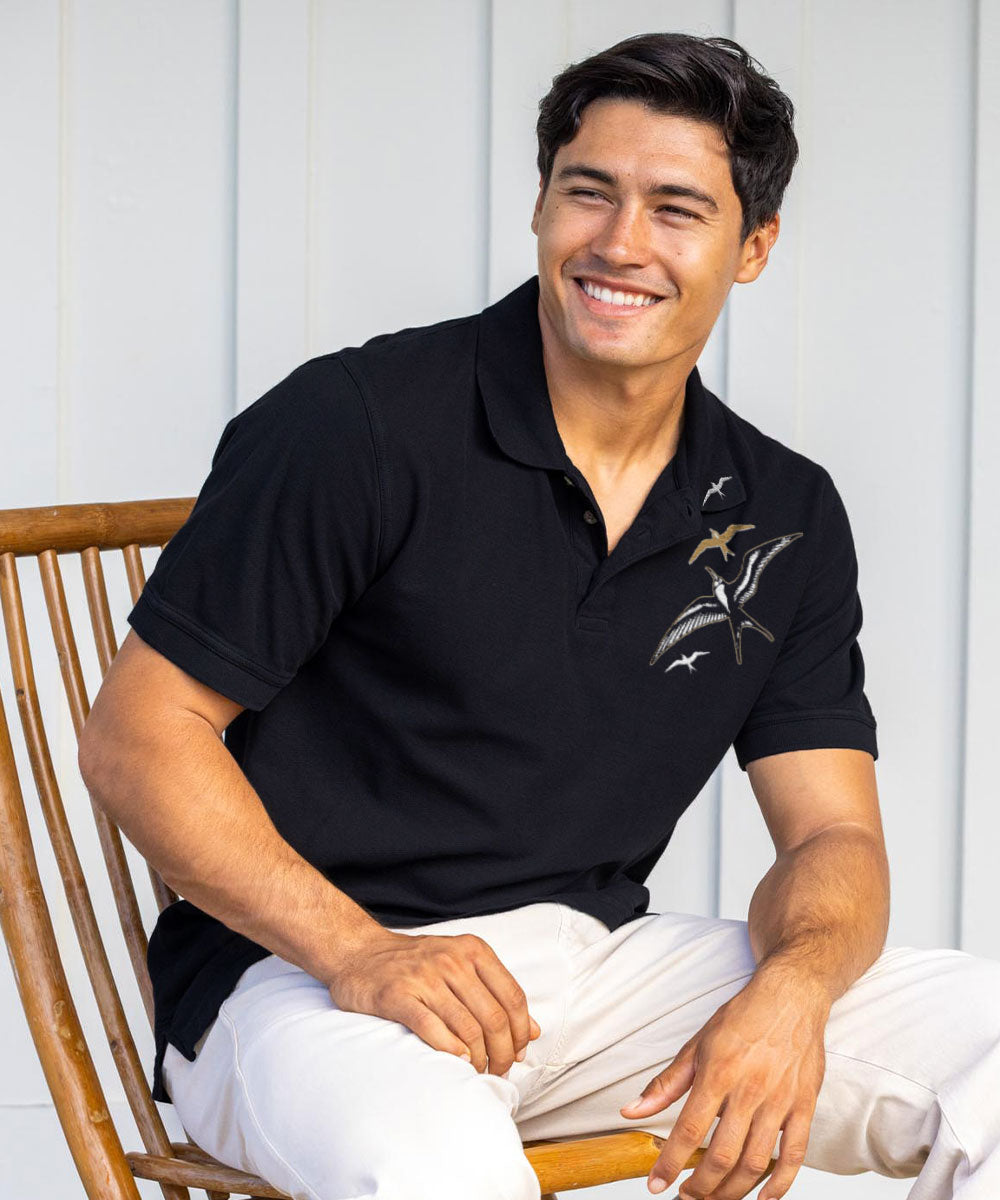 Man wearing a black polo shirt with a logo, sitting on a wooden chair. View 2