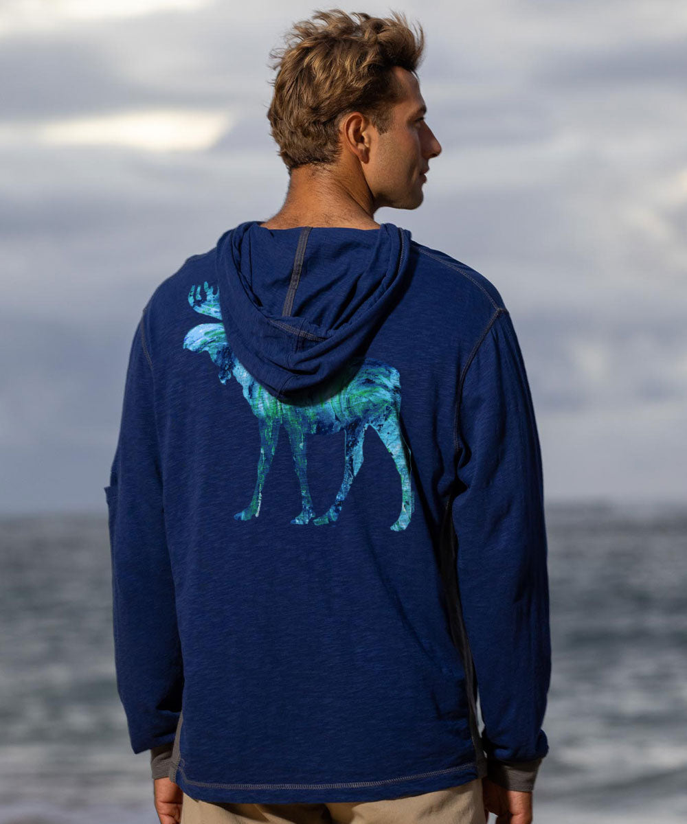 Person wearing a blue hoodie with a graphic of a deer on the back, standing by the ocean. View 2
