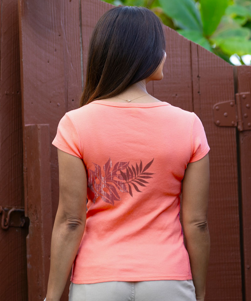 Woman wearing a coral pink t-shirt with a leaf design, standing in front of a wooden gate. View 3