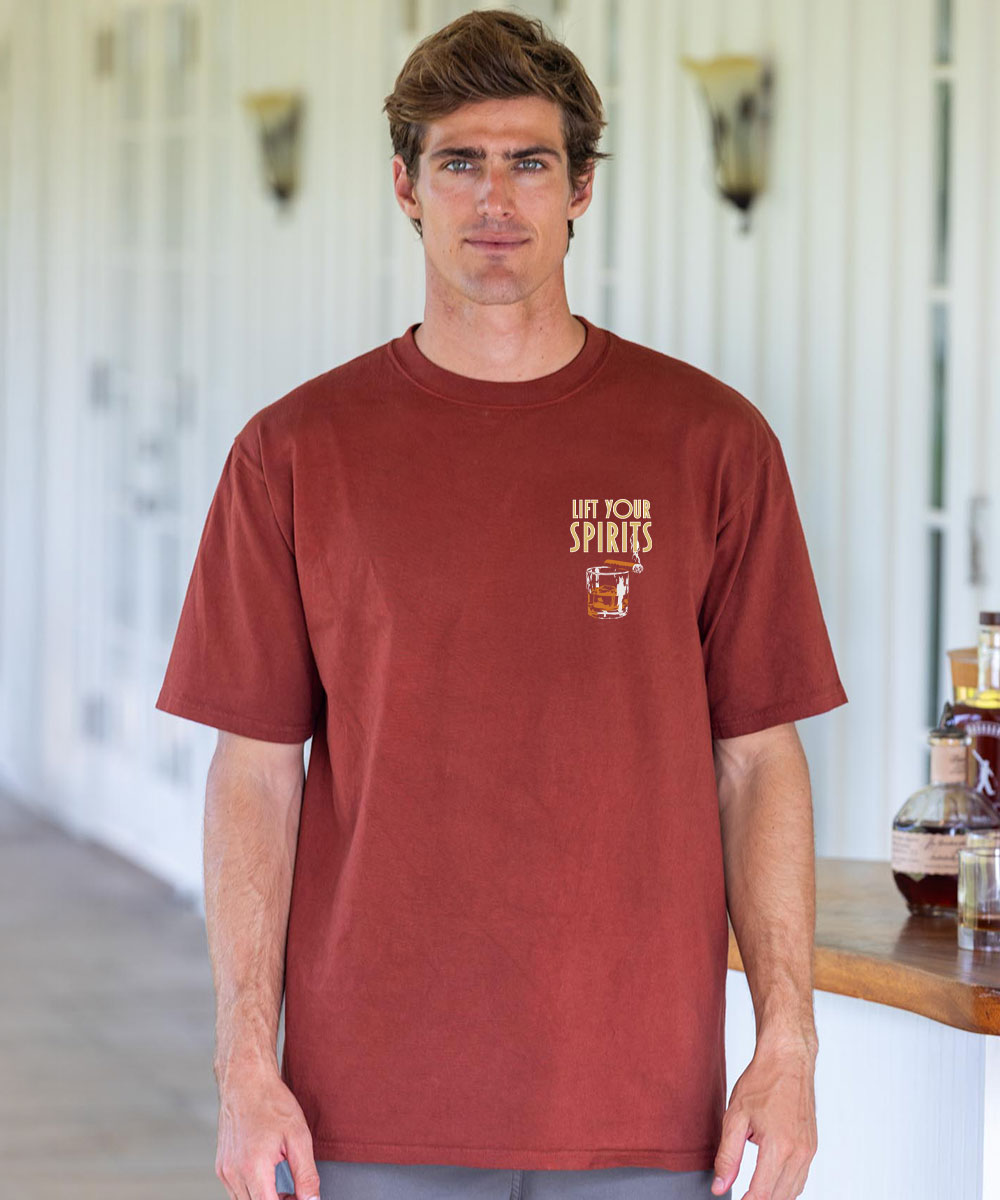 Man wearing a brown t-shirt with a logo and text, standing indoors. View 3