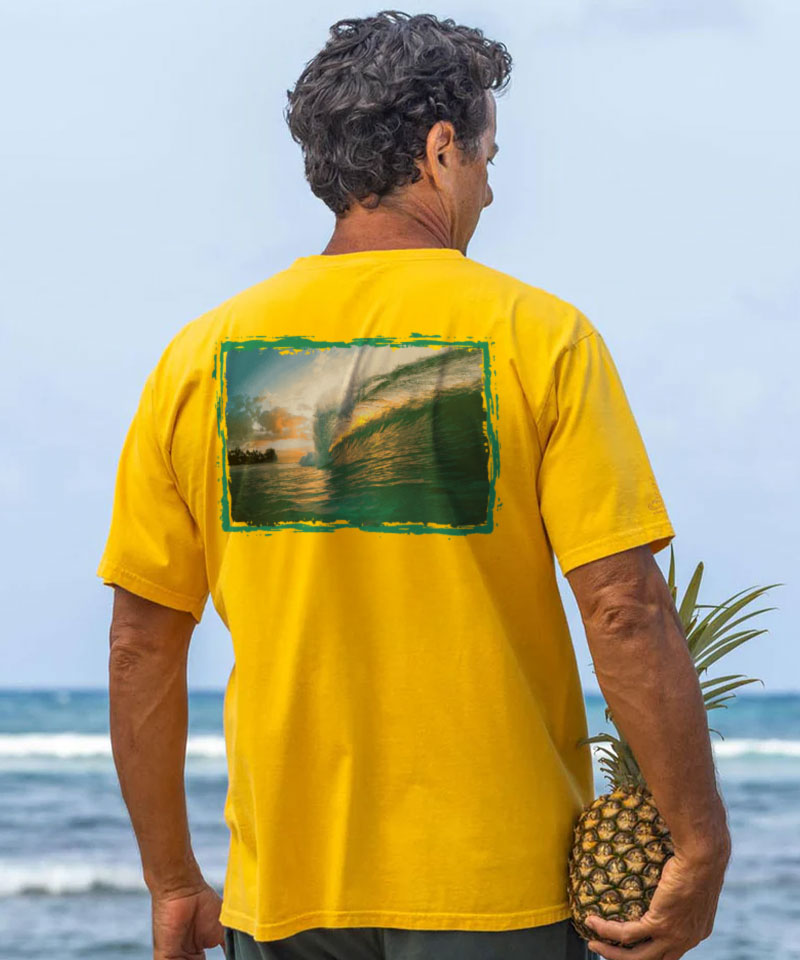Man wearing a yellow t-shirt with a scenic design, holding a pineapple, standing on a beach. View 2